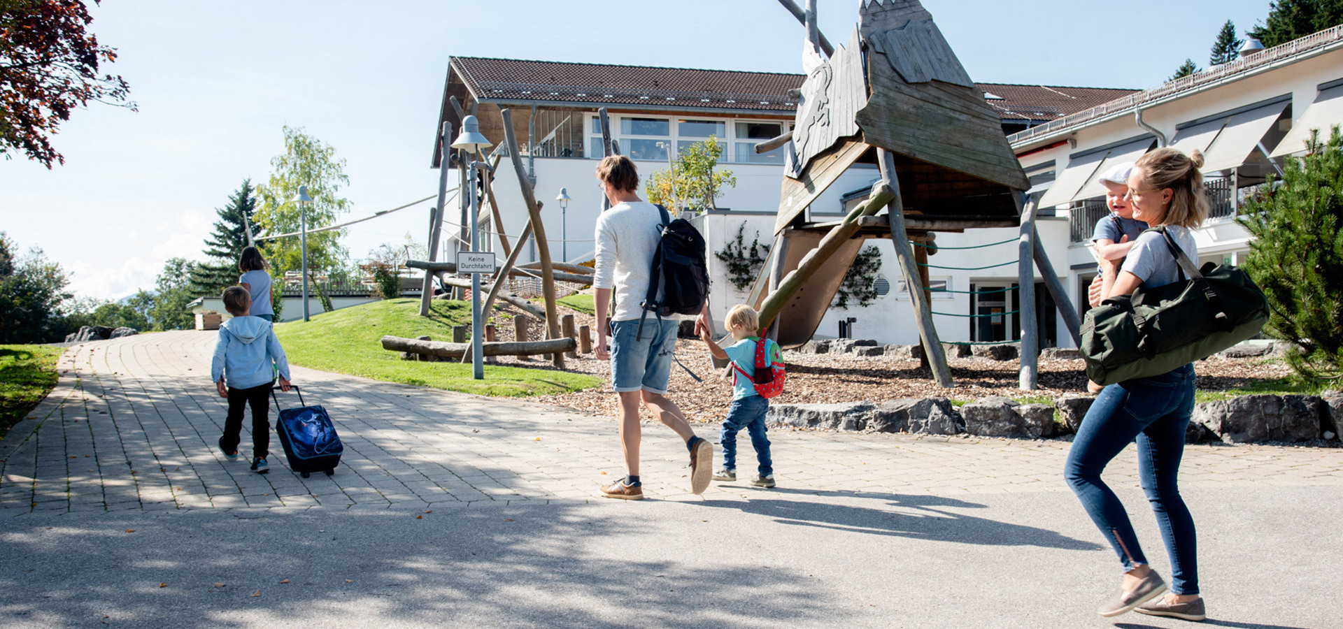 Familie mit vier Kindern auf dem Weg zum Allgäuhaus Wertach , ein Kind und Mann tragen Rucksäcke, eines zieht einen kleinen Koffer, Mutter trägt Baby und Tasche.