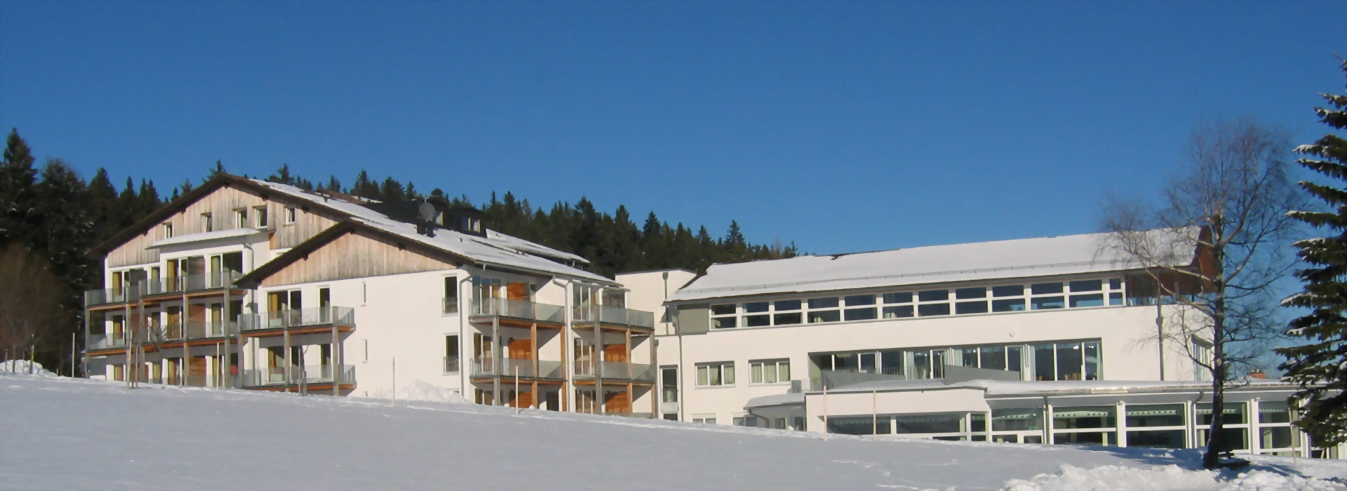 Allgäuhaus Wertach, ein großes weißes Gebäude mit mehreren Balkonen, umgeben von verschneiter Landschaft und Nadelbäumen im Hintergrund.