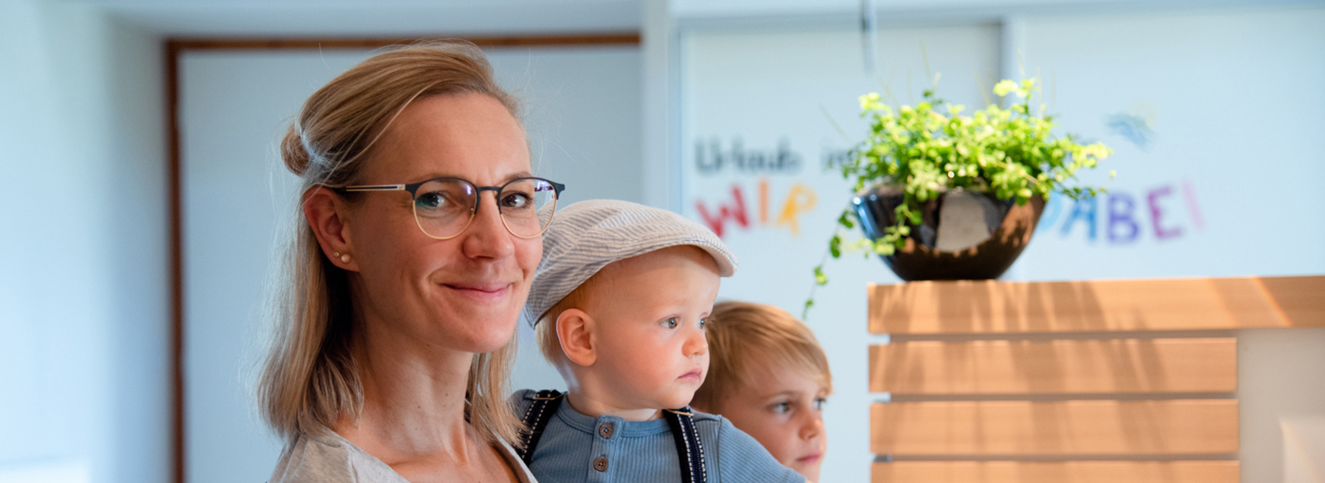 Frau mit Brille hält Kleinkind mit Mütze, im Hintergrund ein älteres Kind und eine Pflanze auf einem Holzregal.