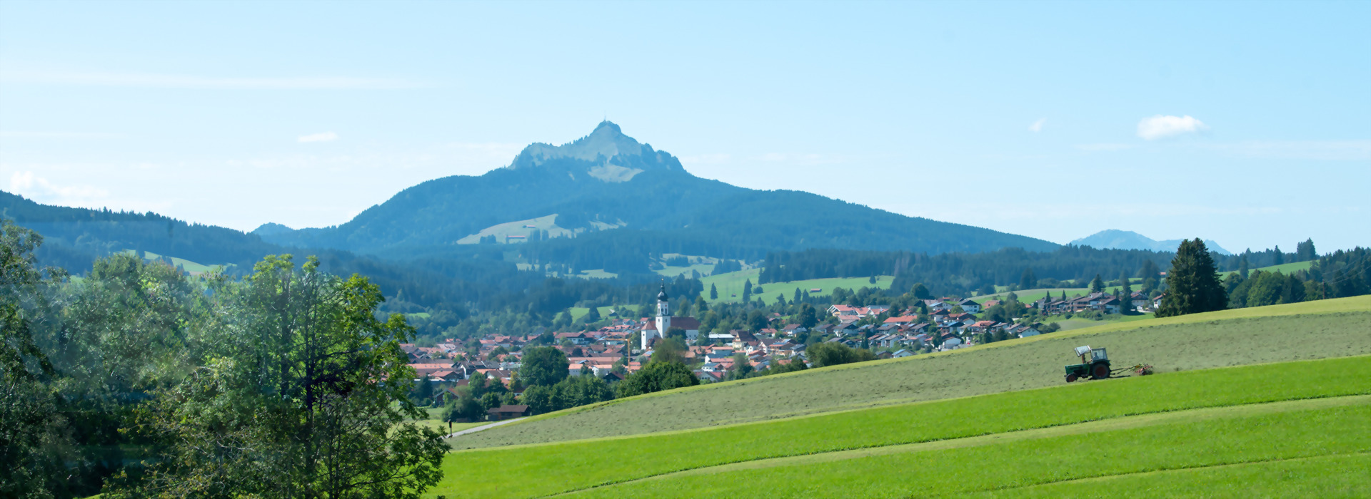 Blick vom Allgäuhaus Wertach auf grüne Wiesen, Dorf mit Kirche und bewaldete Berge im Hintergrund unter blauem Himmel.