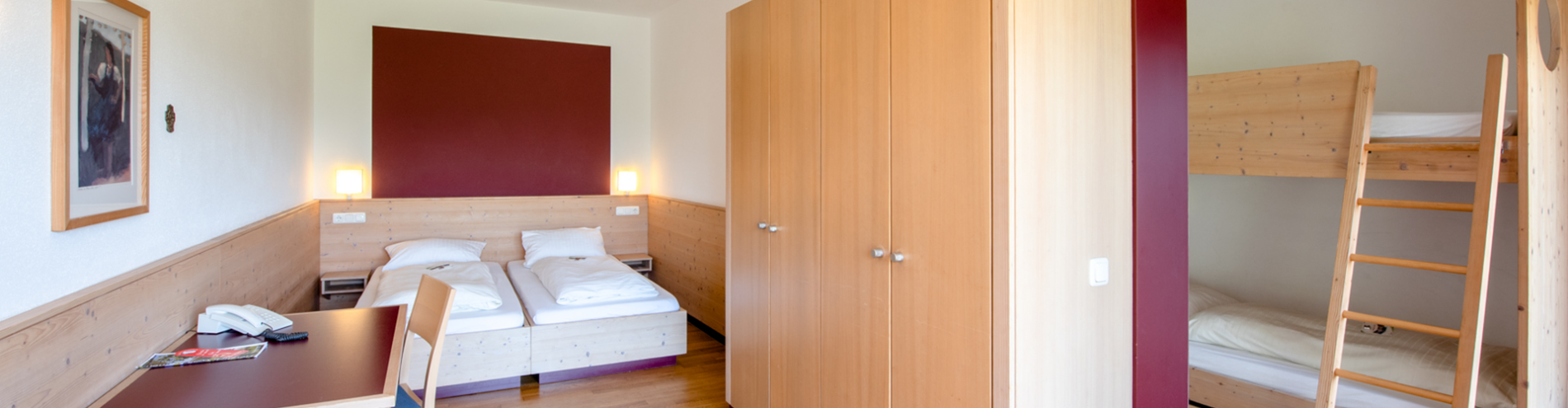 Zimmer mit Doppelbett, rotem Sofa, Schreibtisch mit Stuhl und angrenzendem Raum mit Etagenbett.
