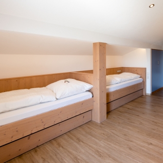 Kinderzimmer vom Allgäu-Appartement mit zwei Einzelbetten aus hellem Holz und rotem Sofa mit Kissen.