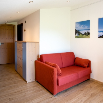 Wohnbereich des Familien-Appartements mit rotem Sofa, Holzboden, zwei Landschaftsfotos an der Wand und einem Holzstuhl.