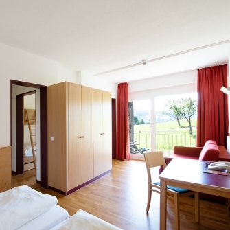 Helles Junior-Appartement mit Doppelbett, Kleiderschrank, Schreibtisch, Blick ins Kinderzimmer und großem bodentiefem Fenster mit Blick auf eine grüne Wiese.