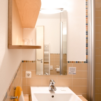 Badezimmer vom Junior-Appartement mit Waschbecken, Spiegel, Handtuchhalter und beige gefliesten Wänden mit buntem Mosaikrand.