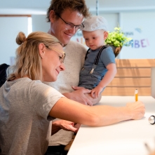 Junge Familie mit Mutter, Vater und Kleinkind an der Rezeption, Mutter schreibt etwas auf Papier, Vater hält Kind auf dem Arm.