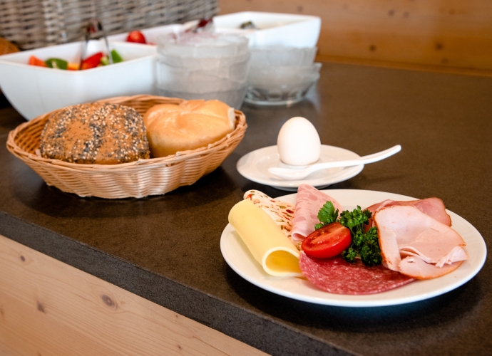 Frühstück am Buffet mit Körbchen voller Brötchen, Teller mit Aufschnitt, Käse, Tomate und Ei auf schwarzer Theke.