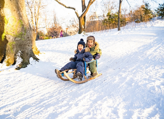 Zwei Kinder in Winterkleidung fahren lachend auf einem Schlitten einen schneebedeckten Hügel hinunter.