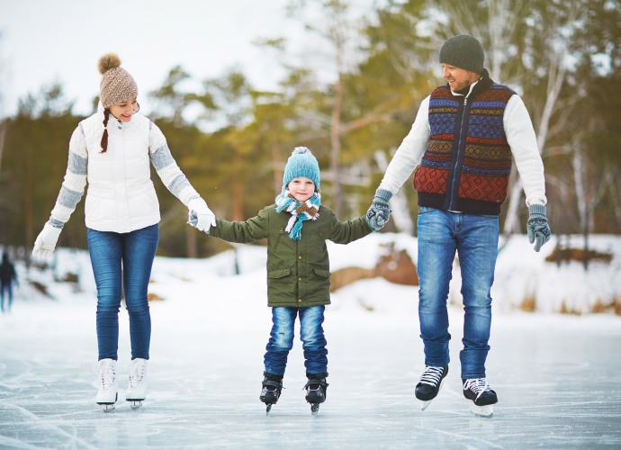 Familie mit Kind hält sich beim Schlittschuhlaufen auf einem zugefrorenen See an den Händen, alle tragen Winterkleidung und Mützen.