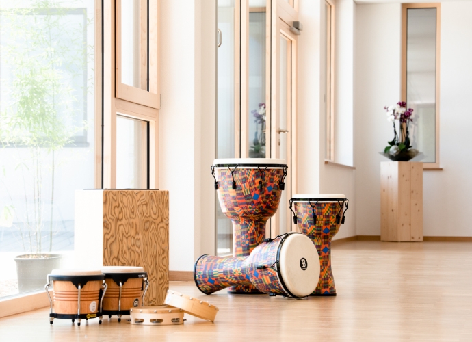 Verschiedene Schlaginstrumente wie Bongos, Tamburin, Cajón und bunte Djemben in einem hellen Musikraum mit großen Fenstern und Holzboden.
