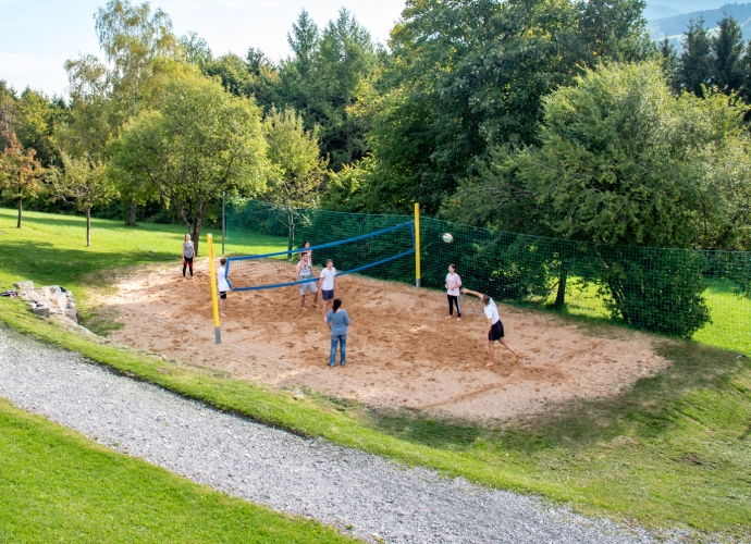 Acht Personen spielen Beachvolleyball auf einem Sandplatz, umgeben von grünen Bäumen und einer Wiese unter blauem Himmel.