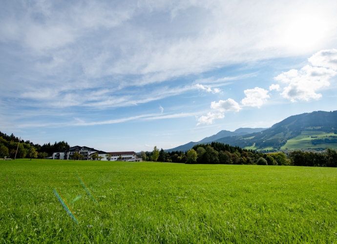 Grüne Wiese mit Allgäuhaus Wertach im Hintergrund, bewaldeten Hügeln und Bergen unter blauem Himmel mit Wolken.