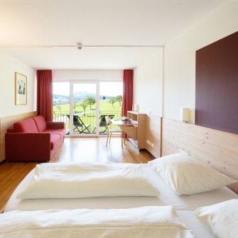 Familien-Appartement mit Doppelbett, rotem Sofa, Schreibtisch und Balkon mit Blick auf grüne Landschaft.