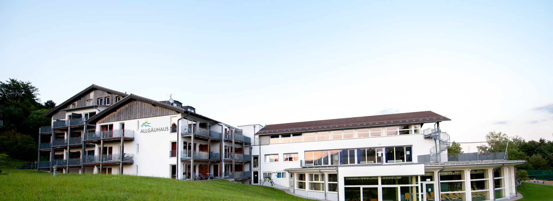 Allgäuhaus Wertach, ein mehrstöckiges Gebäude mit Balkonen und weißer Fassade, umgeben von grüner Wiese und Bäumen.